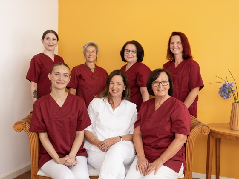 Das Team der Praxis: sieben Mitarbeiterinnen vor der gelben Wand des Behandlungsraums